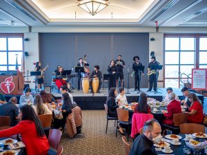 A band with bongos, brass and string instruments performs in front of a large audience at a luncheon