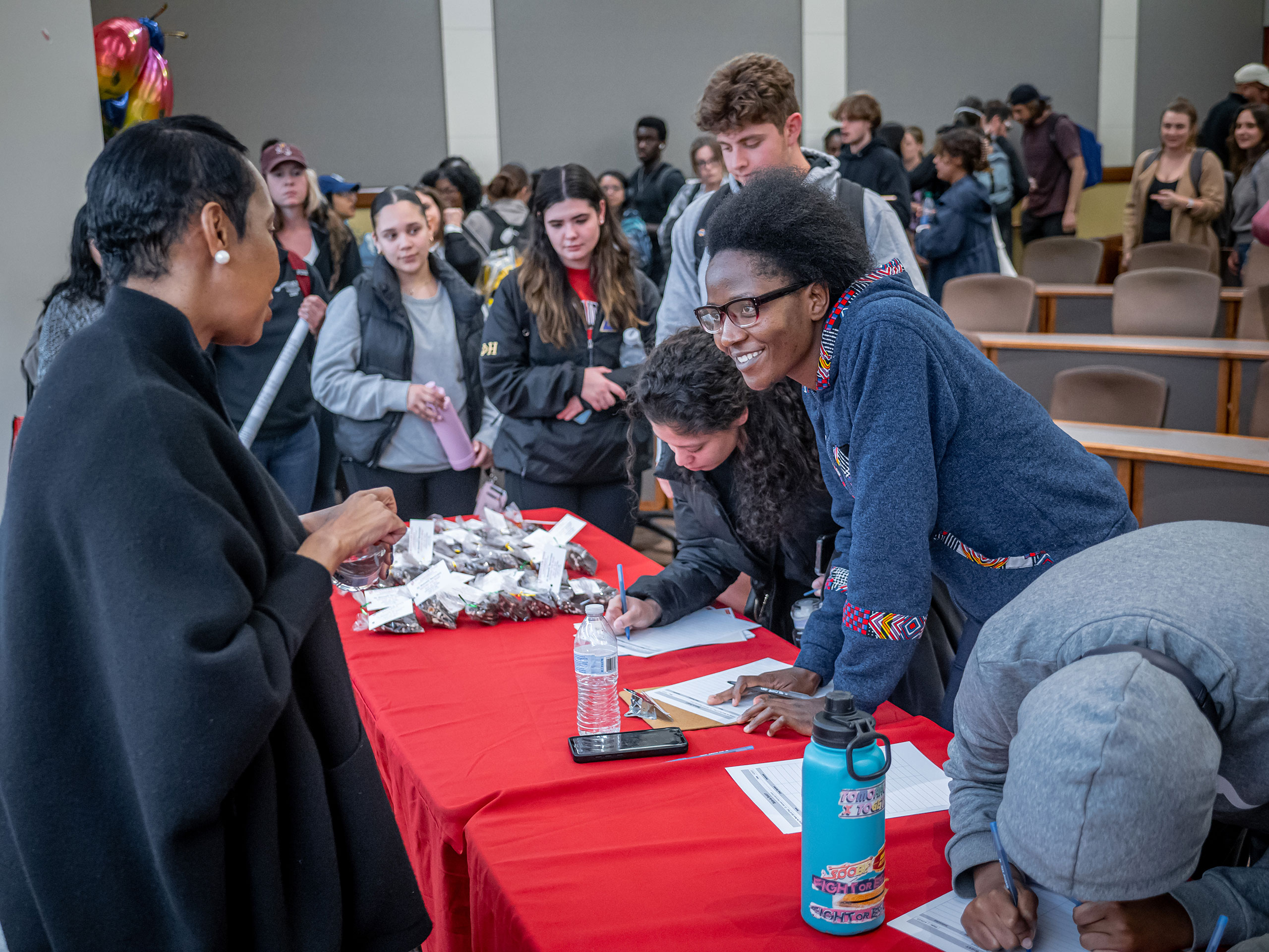 A student smiles and leans forward to talk with a woman, as other students wait their turn in line.