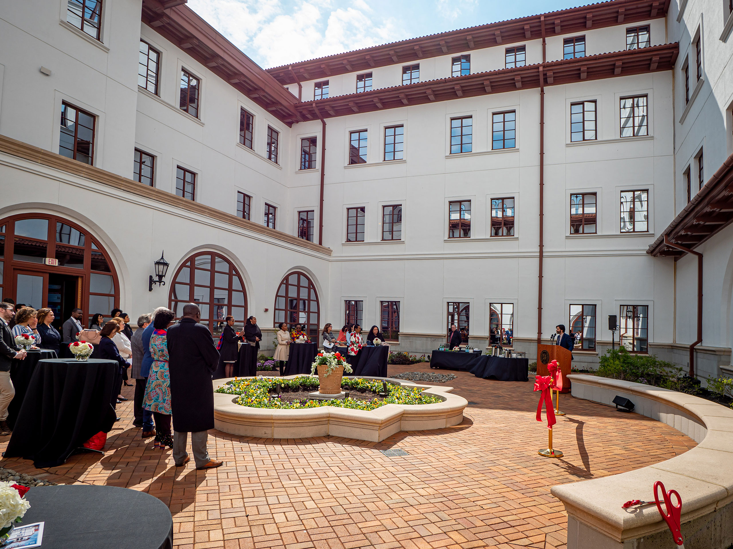 On a sunny day, people stand in a courtyard, listening to a man speaking at a podium.