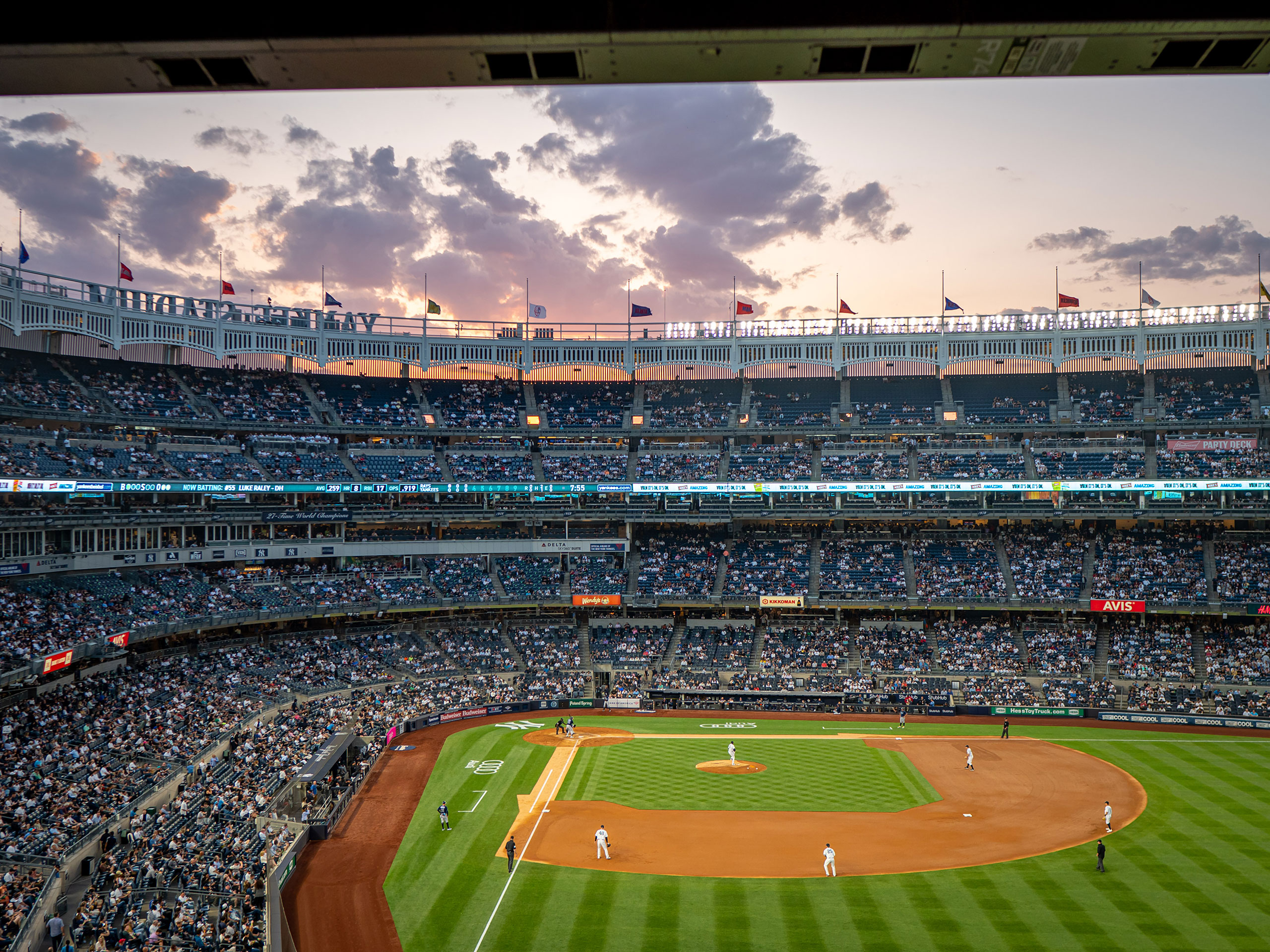 A view of a baseball stadium from box seat level: green field with players in position, a full stadium and a pink and purple sky.