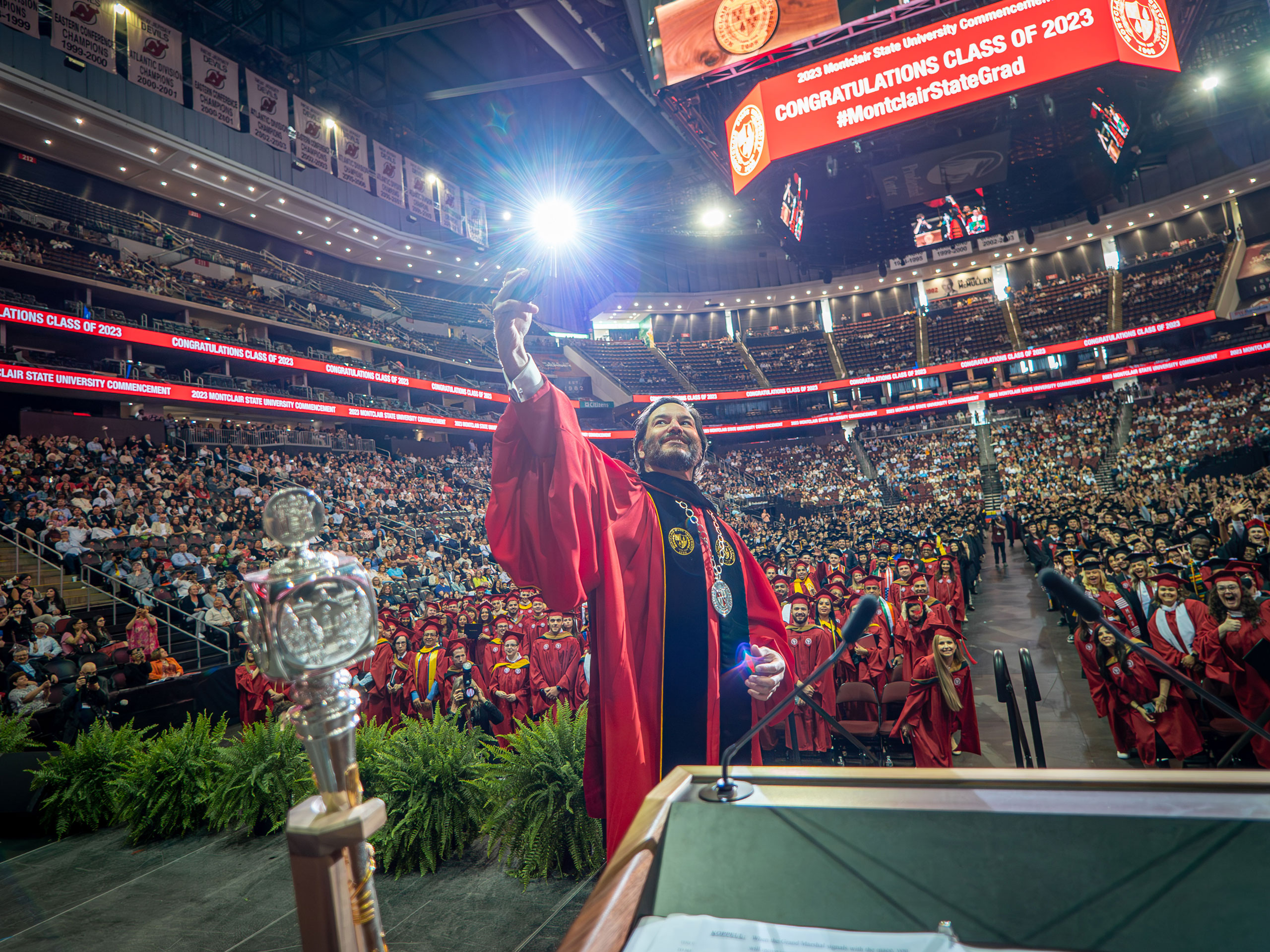 university president in collegiate robes taking a selfie on stage with arena full of graduating students in the background