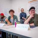 Three people sit at a table and one leans against a wall as they listen to a workshop presentation.