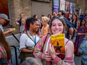 Student holds a playbill from the musical Hamilton near the stage door on a crowded New York City street.