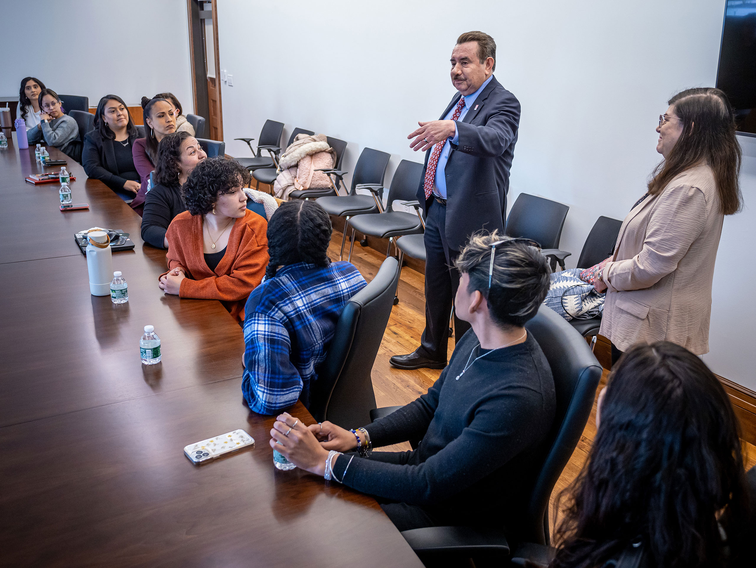 A man gestures as a woman stands next to him in a room filled with people seated at a large conference table.
