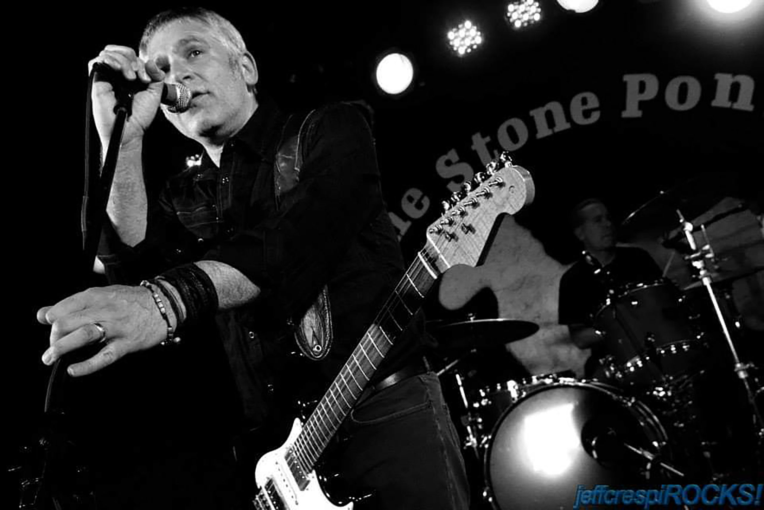 A man with a guitar sings into the microphone with a drummer behind him in front of the words "The Stone Pony."