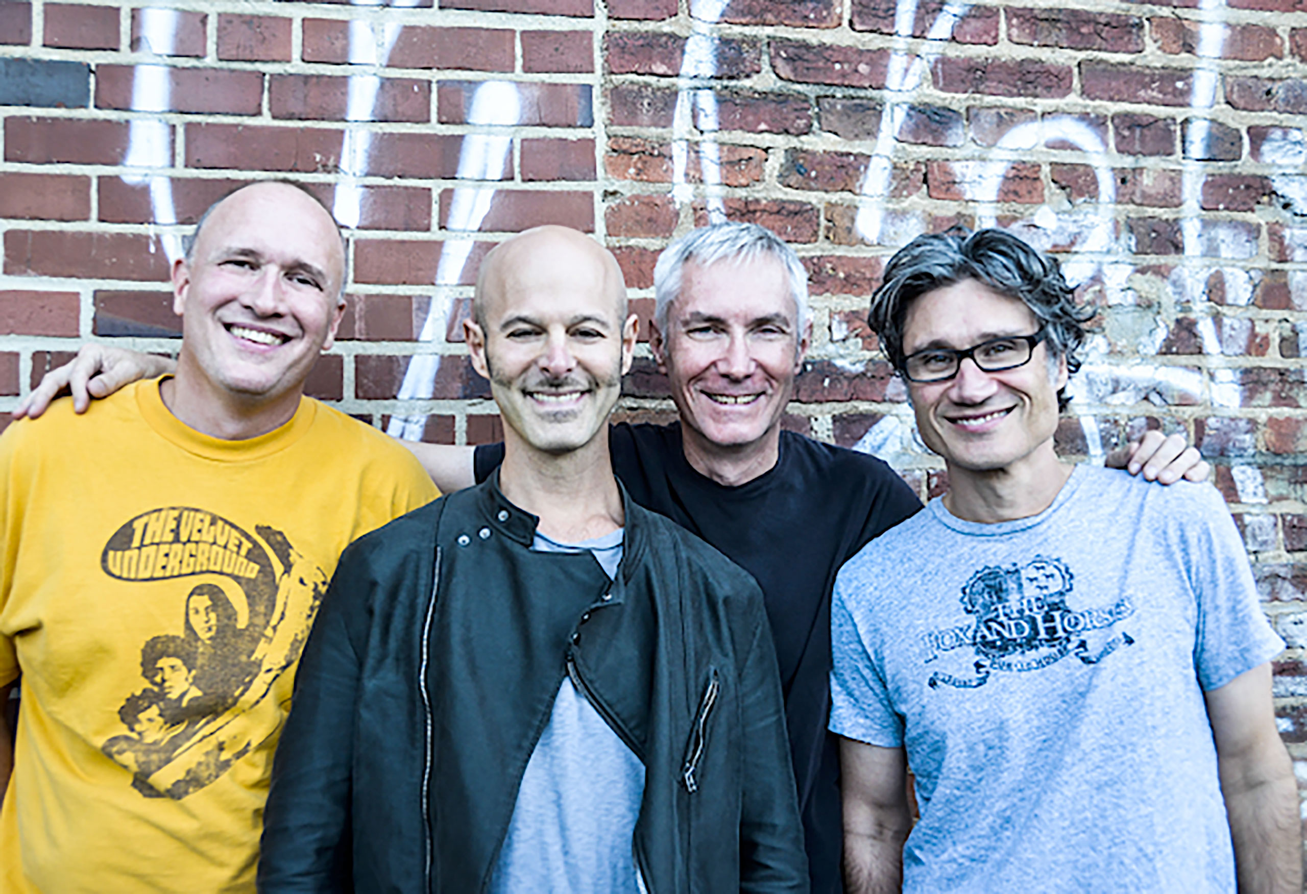Four older men in the band Shudder in 2013 stand in front of a brick wall with graffiti on it.
