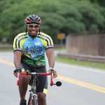 A smiling man in cycling attire that reads Anchor House rides a bicycle.
