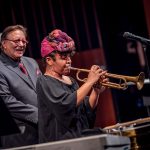 A woman wearing a colorful headscarf plays trumpet as Arturo Sandoval smiles behind her.