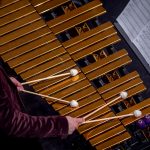 A student in a maroon jacket plays a brass vibraphone with mallets.