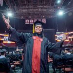 A student in a cap and gown smiles and raises his arms.
