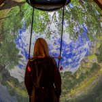 A student wearing a harness stands in the Bertec balance system with a virtual outdoor scene before her.