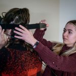 A female student straps virtual reality goggles onto another student.