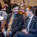 Two men wearing suits chat while seated side by side.