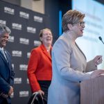 A man and woman laugh while a woman speaks at a lectern.