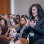 A woman in a black dress stands amid an audience speaking into a microphone.