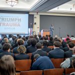 A room filled with young men listen to a speaker on stage.