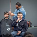 A student speaks into a microphone while another, seated behind him, gets a haircut from a barber.