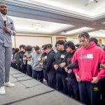 A man stands on a platform with a microphone as men on the floor below lock arms and listen. 