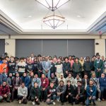 A large group of adults and students pose for a photo onstage.