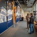 A man with a camera and a microphone interviews another man in front of a line of banners featuring players.