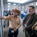  A student wearing a cowboy hat reviews his poster as two people look on.