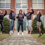 The Povolo quintuplets wearing Montclair State University sweatshirts jump together in front of a high school building.