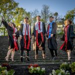 The Povolo quintuplets wearing graduation gowns jump together in front of stone steps outdoors.