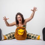 Eliza Andrus laughs and opens her arms wide sitting behind Ricky’s mascot head and wearing oversized sneakers.