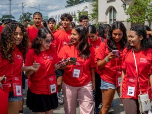 A group of students in red t-shirts and lanyards walk together in a large group on college campus.