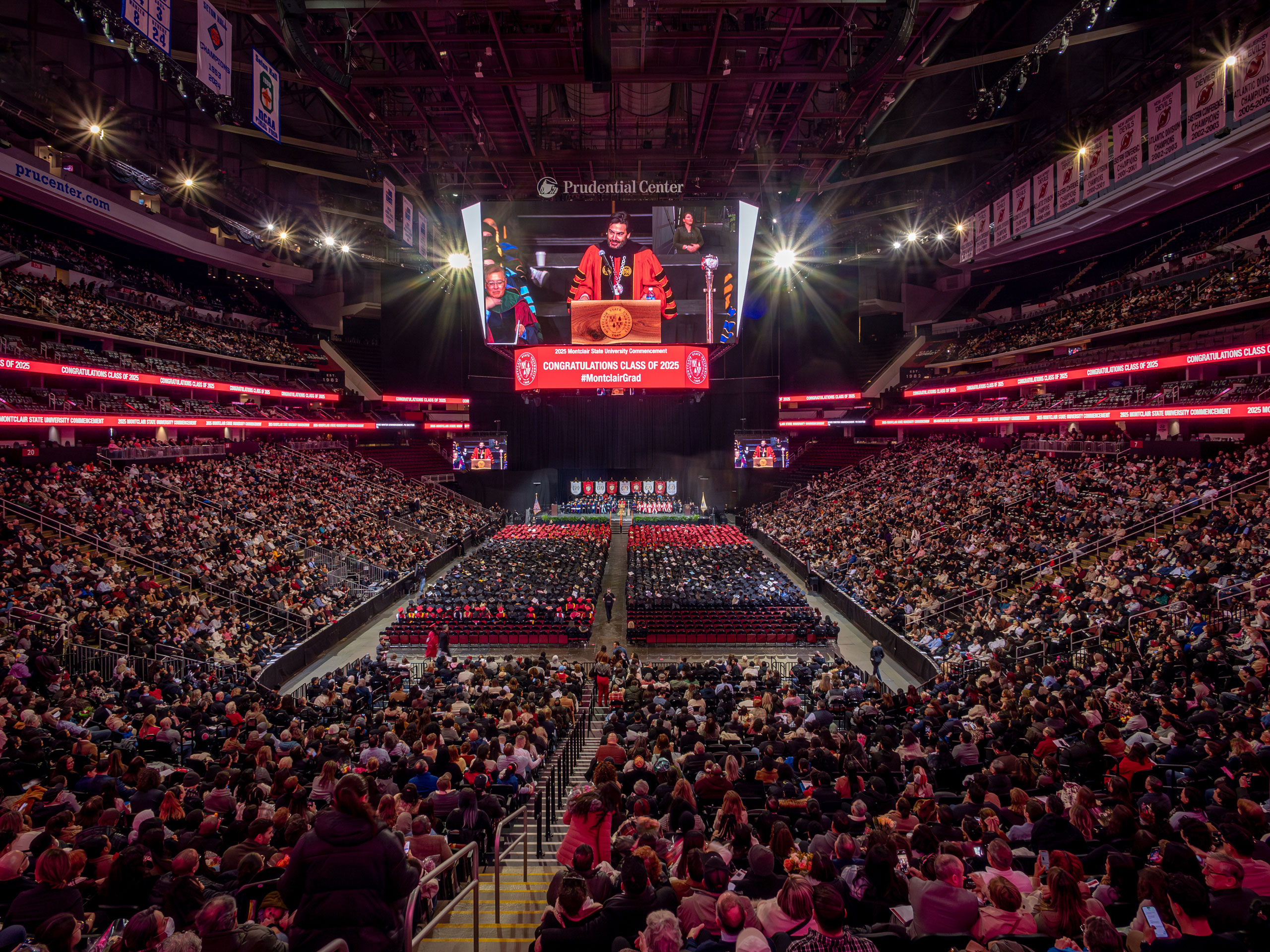 A wide shot of the Prudential Center captures the large crowd in the arena, with graduates seated on the floor in their caps and gowns. Above them, on the jumbo screen, the University president is visible, addressing the audience.