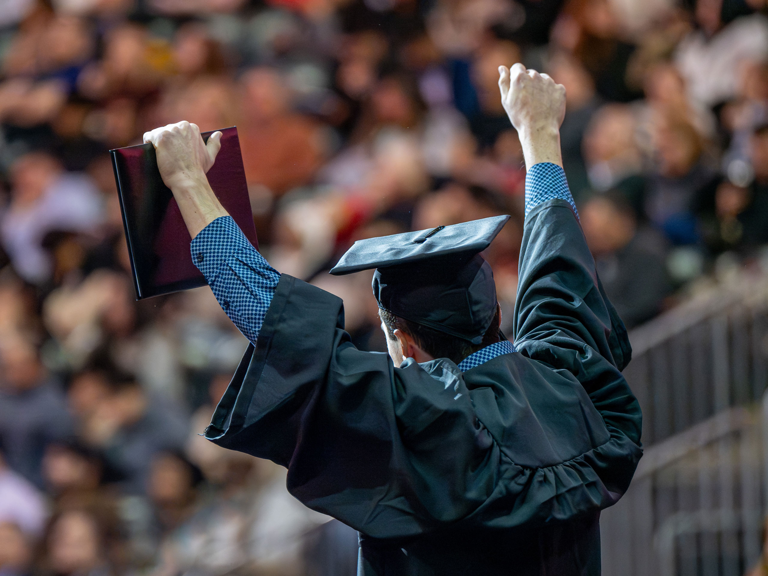 A graduate raises his arms in triumph after receiving his diploma, shown from the back.