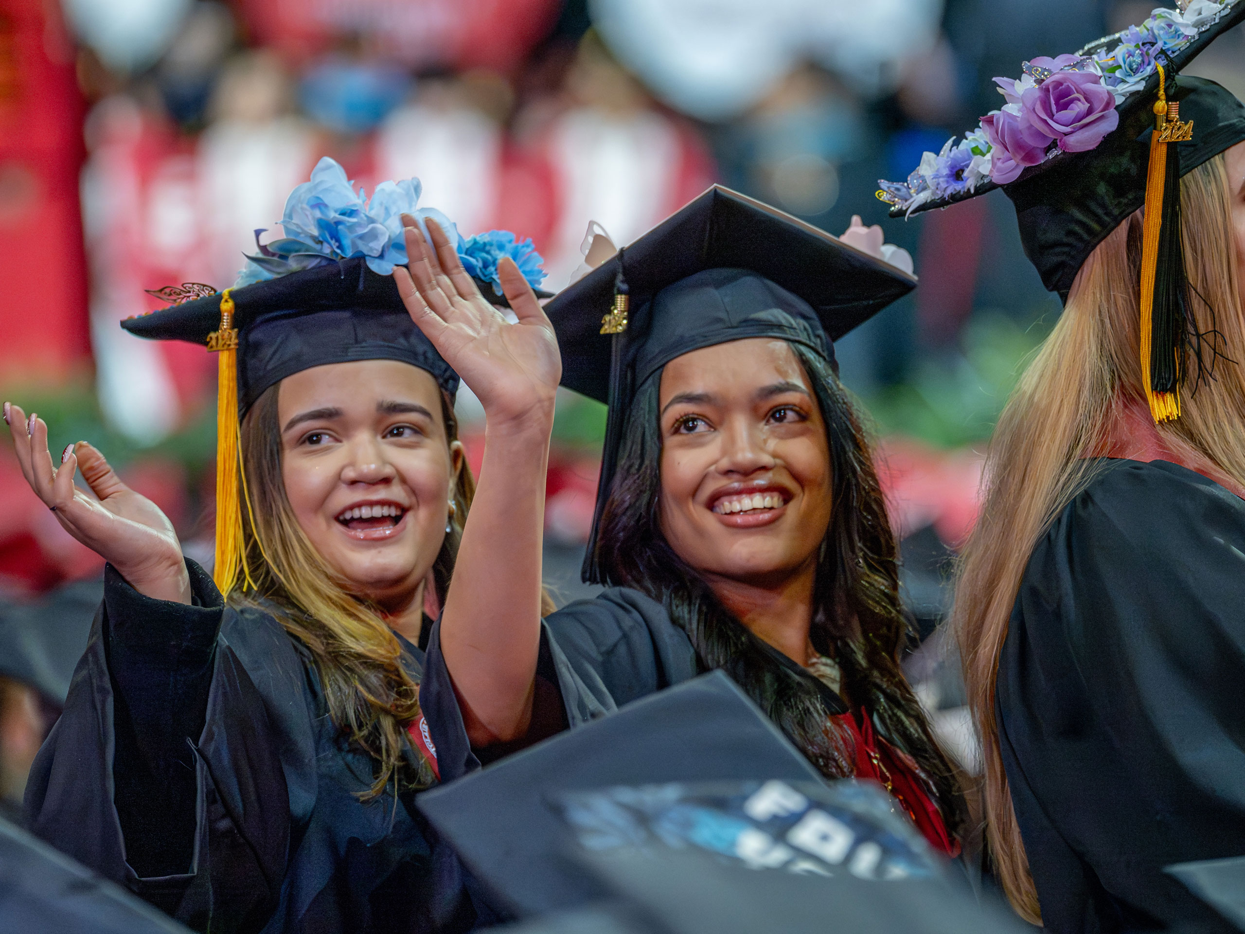 Two graduates, seated on the floor of the Prudential Center, turn and wave. Their caps are decorated with flowers.