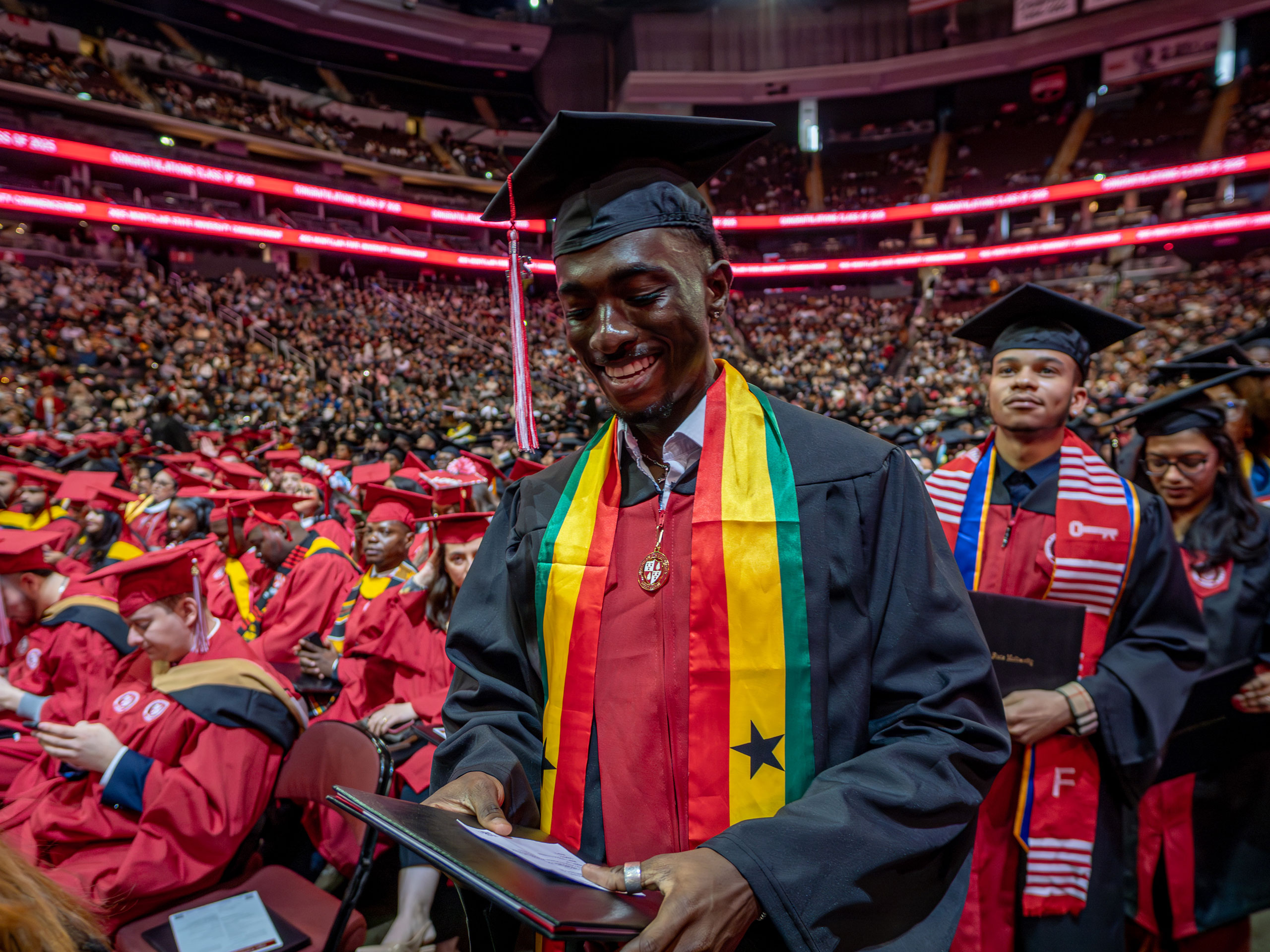 A graduate wearing a stole in red, yellow, and green with a black star proudly looks at their diploma after receiving it.