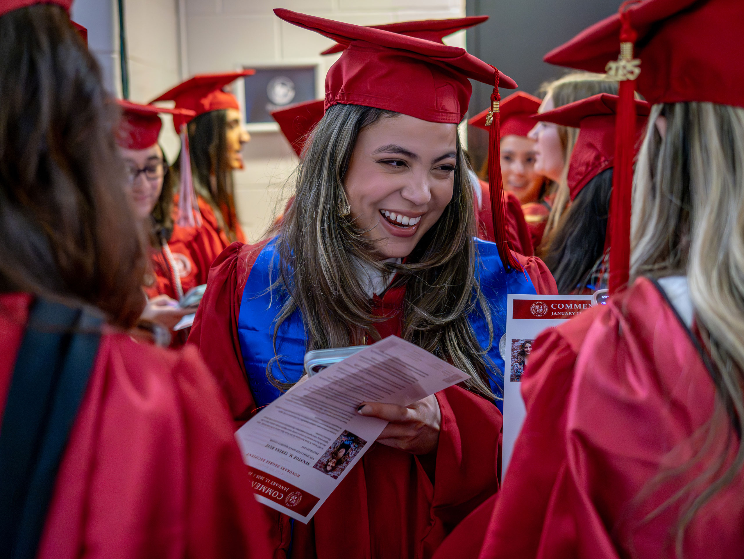 A graduate in a red gown smiles, surrounded by fellow graduates, while holding the day's program.