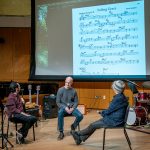 Three men onstage discuss a sheet of music projected overhead on a screen.
