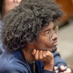 A student wearing glasses holds his hand under his chin in a class.