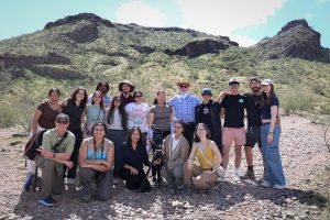 Steve McCarthy and Thomas E. Franklin stand in the Arizona desert with 14 student journalists who worked on Arizona Stories: Border, Water and Politics. The group poses together during their reporting trip, dressed casually with camera equipment visible against a rugged desert landscape.