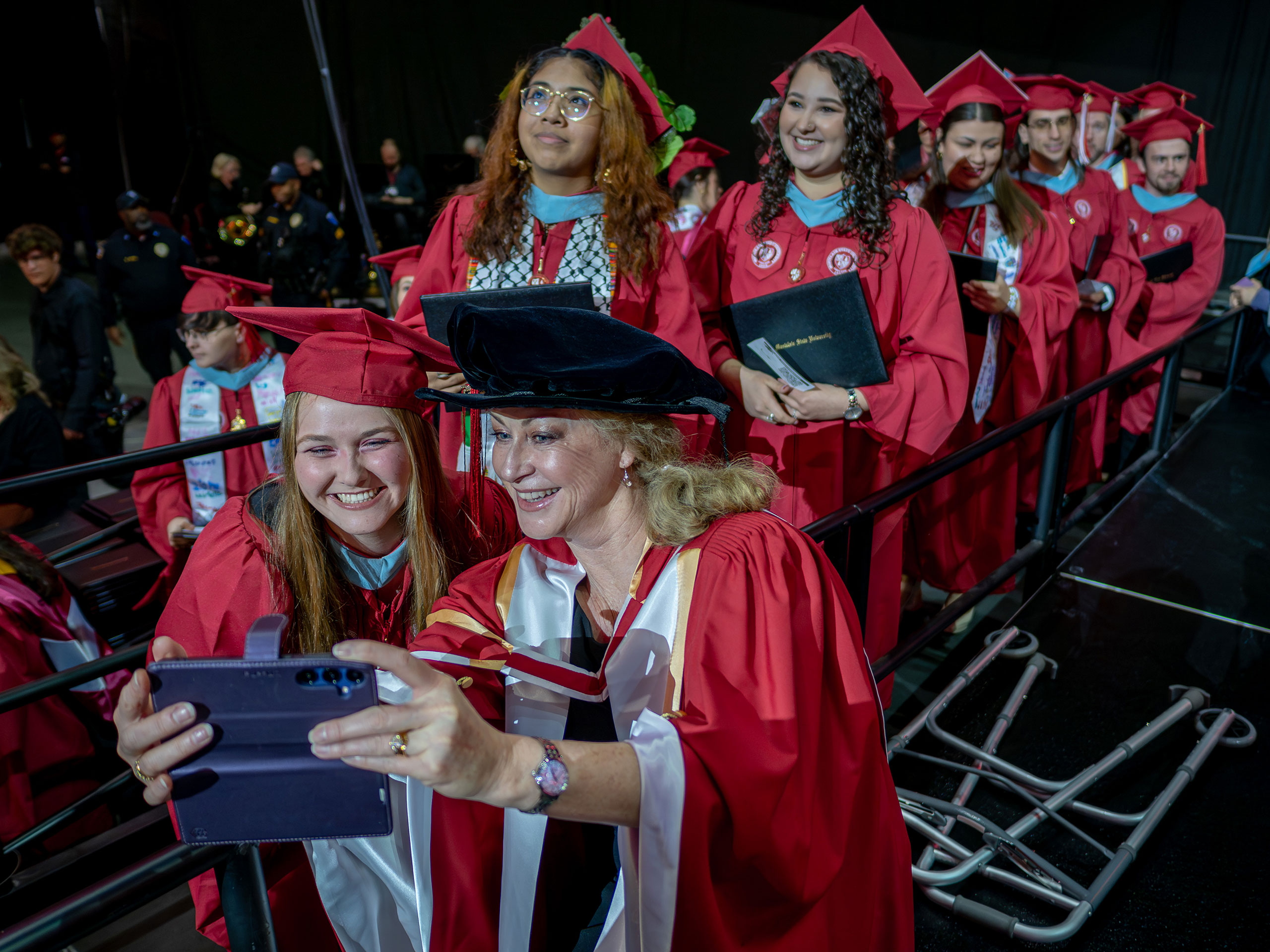 A faculty member in red gown poses for a selfie with a graduate.
