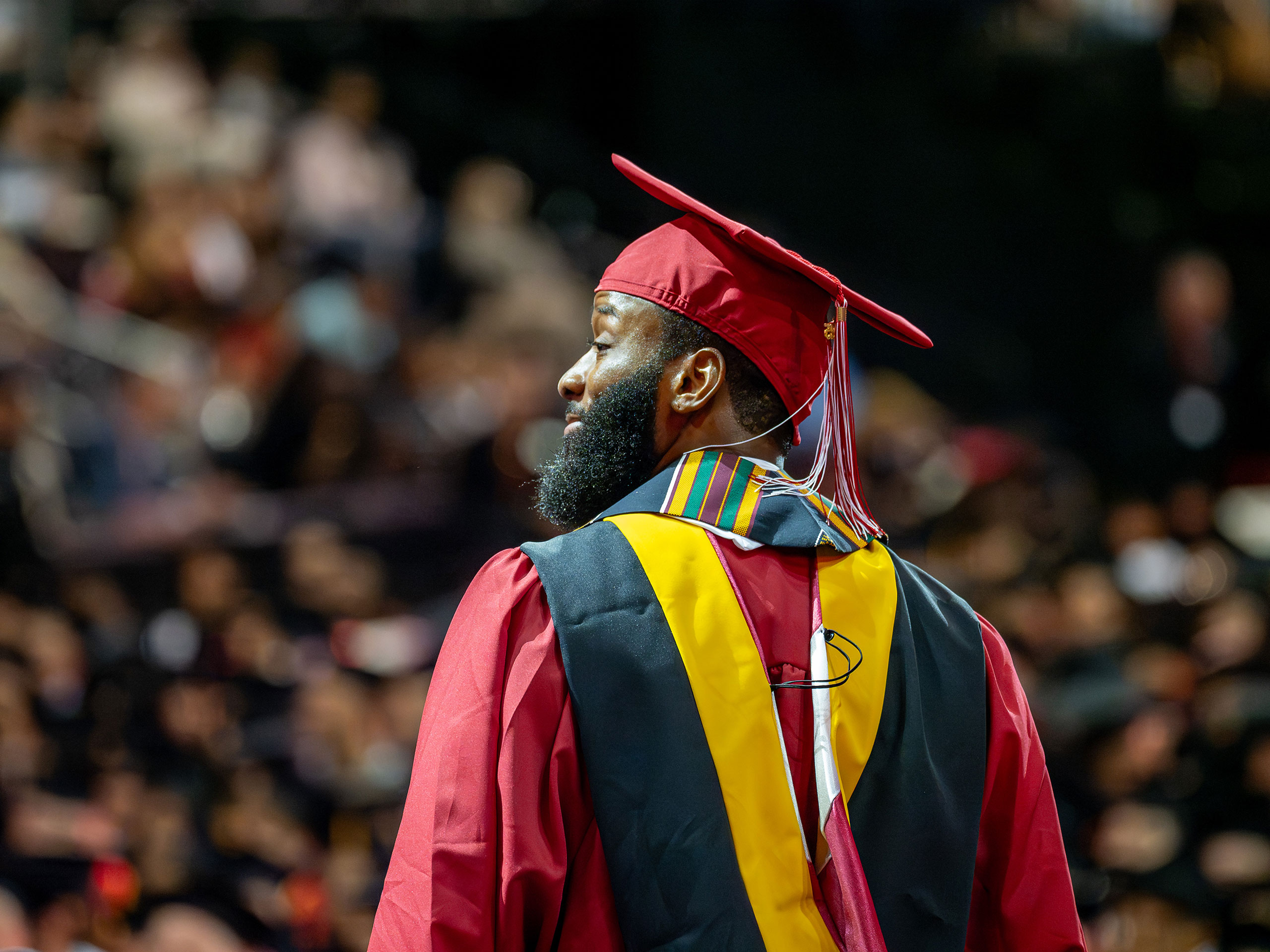 A master's graduate in red cap and gown looks toward the crowd from the stage