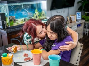 Oscaterin embraces Kamila at the breakfast table, sharing a warm moment to start the day. A plate of eggs, ham and pancakes sits beside Kamila.