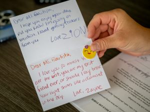 A hand holds open a card filled with handwritten thank-you notes from children, expressing love and appreciation for Ms. Bautista.