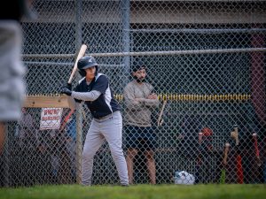 A child wearing a baseball helmet and uniform stands at bat on a baseball field, ready to hit, while an adult watches from behind the fence.