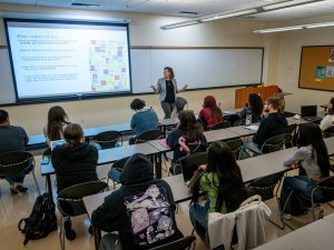 A college classroom with students seated at desks and a professor teaching at the front, a slide projected with class expectations visible on the screen.