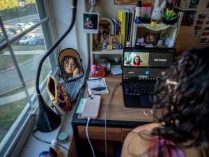 Sara Flores sits at her desk during a Zoom meeting on her laptop. Her reflection appears in a small mirror beside her.