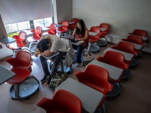 A student naps on a classroom desk while Sara Flores sits nearby, checking emails on her laptop.