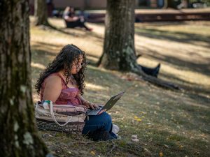 Sara Flores sits under a campus tree, working on her laptop.