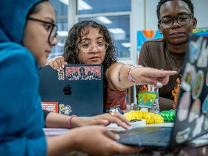 Sara Flores sits in the center between two students, pointing at a laptop screen while explaining how to find training opportunities and record volunteer hours.
