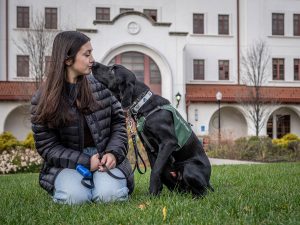 Sara Brocken sits on grass outside the Feliciano School of Business, nuzzling affectionately with Fuji, a young black Labrador wearing a green “Seeing Eye Puppy” vest.