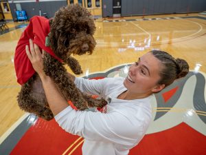 An athlete in a white shirt stands on a basketball court, holding Pebbles, a mini chocolate labradoodle, dressed in a red shirt.