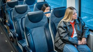 Emma Haskell sits alone on a seat inside a commuter train. Several other passengers sit in adjacent rows, as the train travels along its route.